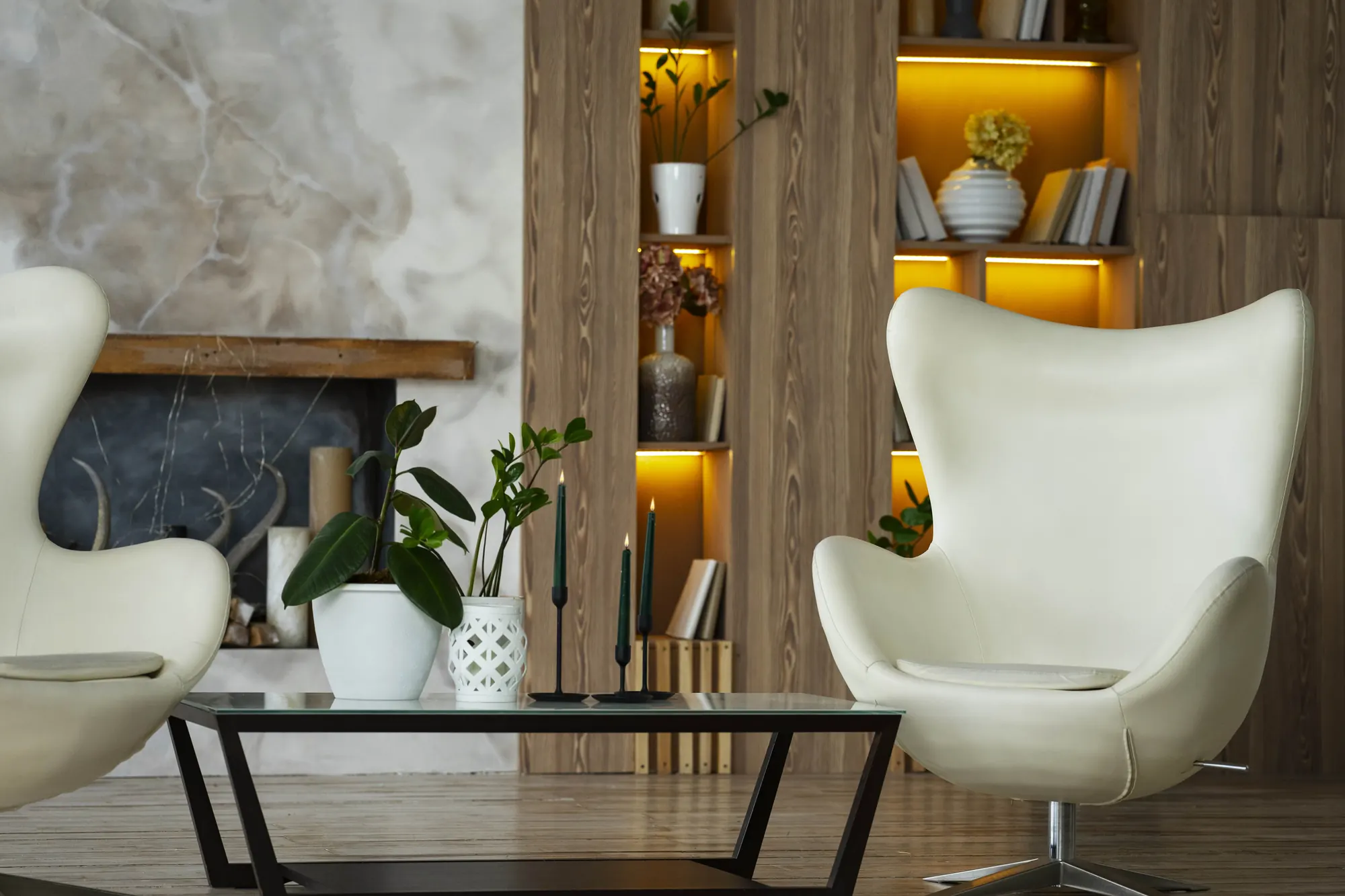 Coin salon : deux fauteuils coque blancs, table basse à plateau de verre, bibliothèque en bois à niches rétroéclairées.