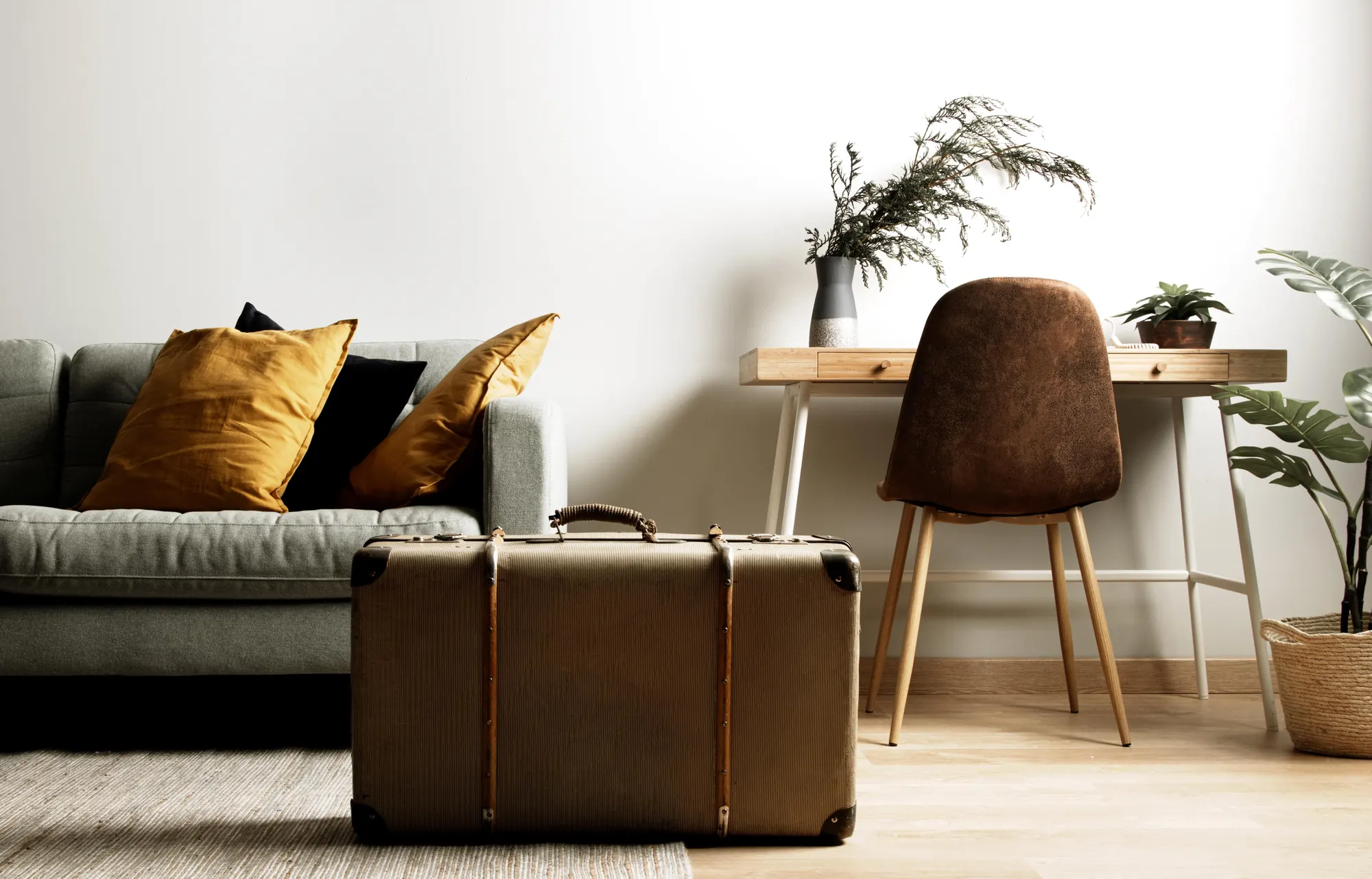 Séjour clair : canapé gris à coussins ocre, valise en cuir posée au sol, table et chaise en bois devant un mur blanc.