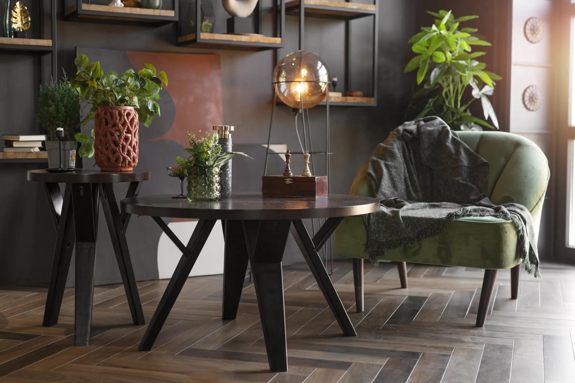 Salle à manger sombre : table en bois sur piètement noir, parquet à chevrons, étagères murales garnies de plantes.