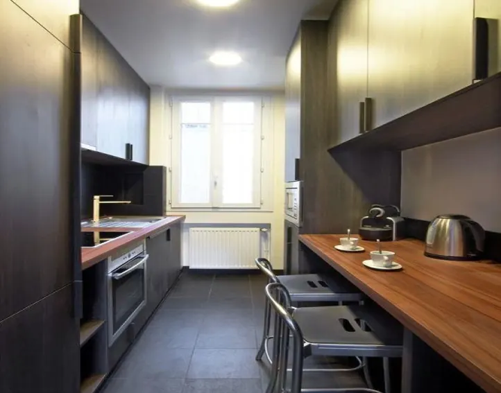 Cuisine en longueur : façades sombres, plan de travail en bois, tabourets et fenêtre en fond.