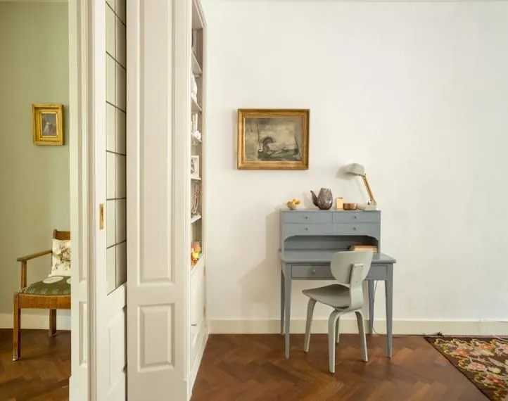 Pièce ancienne à moulures : parquet à chevrons, petit bureau peint et tableaux encadrés.