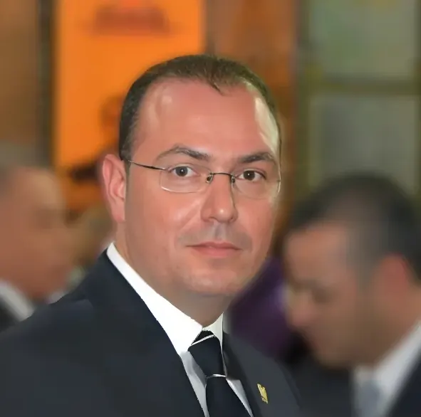 Portrait d’un homme en costume sombre et cravate, lunettes fines, devant des panneaux orangés.